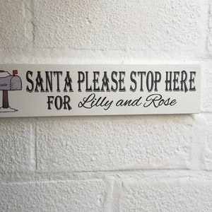 Könnte beinhalten: Ein weißes Holzschild mit schwarzem Text, der "SANTA PLEASE STOP HERE FOR Lilly and Rose" lautet. Auf der linken Seite des Schildes befindet sich ein Cartoon-Bild eines Briefkastens mit einem Weihnachtsmann-Hut.