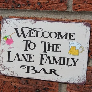Peut inclure: Panneau métallique rectangulaire avec l'inscription "Welcome To The Lane Family Bar" en lettres noires. Le panneau a un aspect vieilli et vintage et présente des illustrations d'un cocktail et d'une chope de bière.