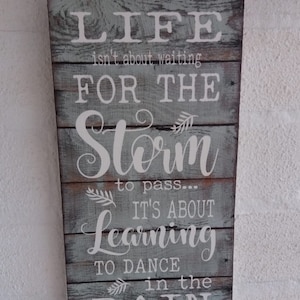 May include: A wooden sign with a distressed, light blue paint finish. The sign features a white inscription that reads: "Life isn't about waiting for the storm to pass... It's about learning to dance in the rain."