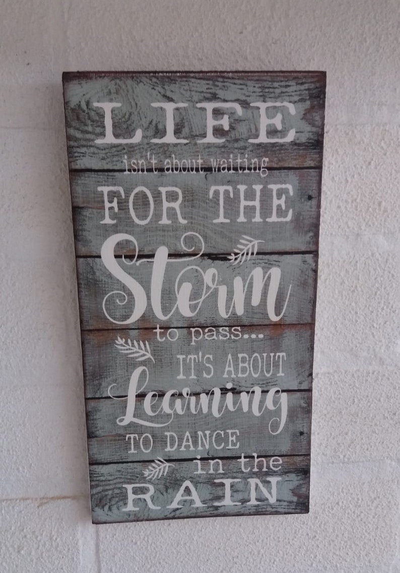May include: A wooden sign with a distressed, light blue paint finish. The sign features a white inscription that reads: "Life isn't about waiting for the storm to pass... It's about learning to dance in the rain."
