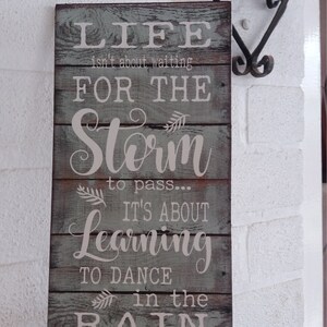 May include: A wooden sign with a distressed, weathered look. The sign has a light blue background with white lettering. The text reads: "Life isn't about waiting for the storm to pass... It's about learning to dance in the rain."