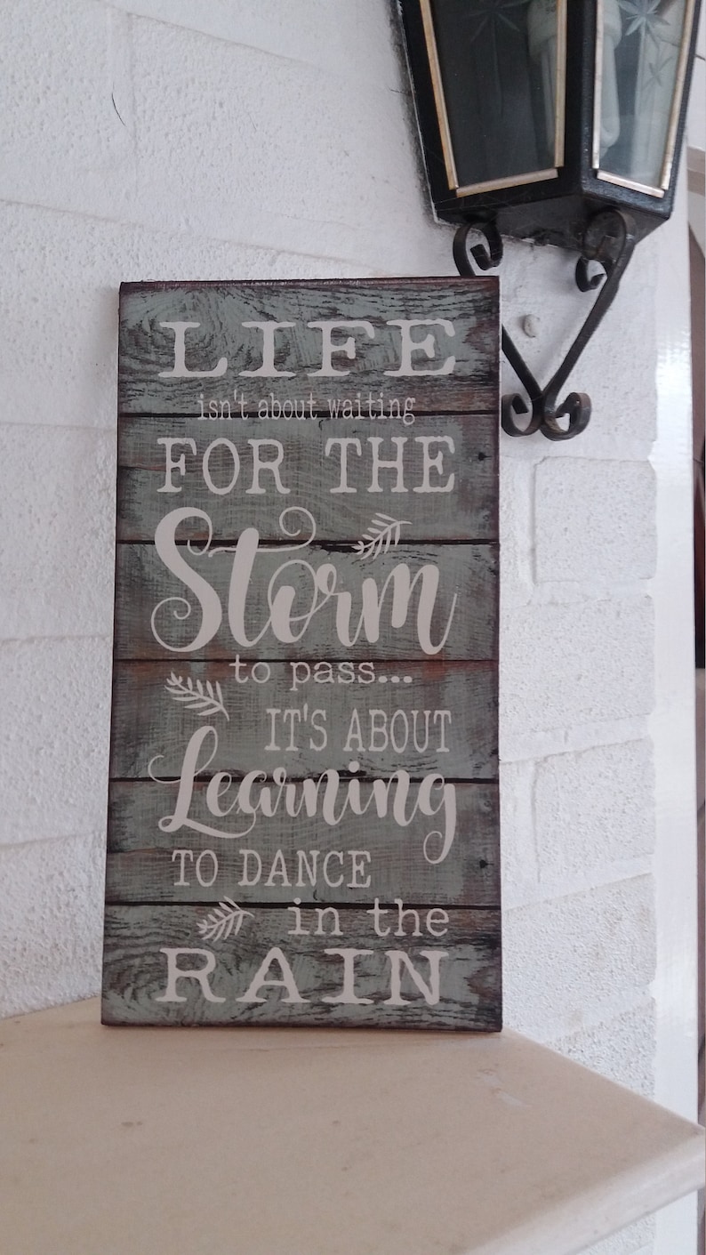 May include: A wooden sign with a distressed, weathered look. The sign has a light blue background with white lettering. The text reads: "Life isn't about waiting for the storm to pass... It's about learning to dance in the rain."