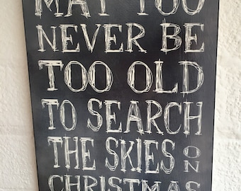 May include: A black wooden sign with white lettering that reads "MAY YOU NEVER BE TOO OLD TO SEARCH THE SKIES ON CHRISTMAS EVE". The sign has a snowflake design at the top and bottom.