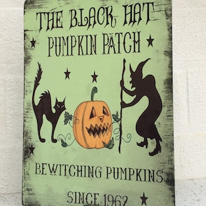 Puede incluir: Un letrero verde con el texto "The Black Hat Pumpkin Patch" y "Bewitching Pumpkins Since 1962". El letrero presenta un gato negro, una calabaza tallada y una silueta de bruja.
