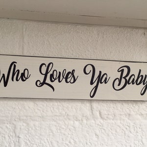 May include: A rectangular, cream-coloured wooden sign with the words "Who Loves Ya Baby" in black cursive script. The sign is mounted on a white brick wall, suitable for home decor or a similar setting.
