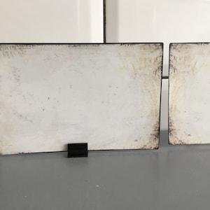 May include: Two rectangular white signs with black borders and black stands. The signs have a distressed, aged appearance with brown staining around the edges. The signs are displayed on a grey surface.