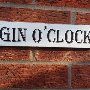 Könnte beinhalten: Ein rechteckiges, weißes Holzschild mit dem Schriftzug "GIN O'CLOCK" in schwarzer, dekorativer Schrift. Das Schild ist an einer roten Backsteinmauer befestigt und erzeugt eine rustikale Ästhetik. Ideal für eine Hausbar oder Küche.