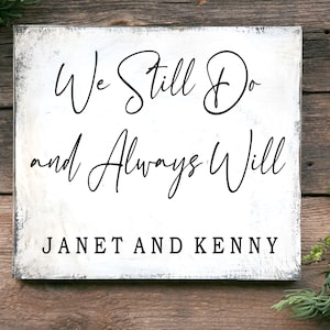 Può includere: Un cartello quadrato in legno bianco con testo nero che recita "We Still Do and Always Will" e i nomi "JANET AND KENNY" in basso. Il cartello ha un aspetto invecchiato e rustico con sfondo bianco e bordo scuro.