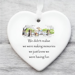 Op de afbeelding: Wit hartvormig keramisch ornament met een lint om op te hangen. Het ornament heeft een kleurrijke illustratie van een beer en een jongen bij een houten hek. De tekst luidt: "We realiseerden ons niet dat we herinneringen aan het maken waren, we wisten gewoon dat we plezier hadden."