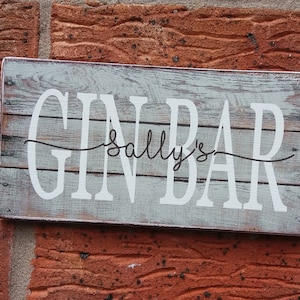 May include: A white wooden sign with the text "GIN BAR" in black lettering and "Sally's" written in cursive above the word "BAR". The sign has a distressed, weathered look.