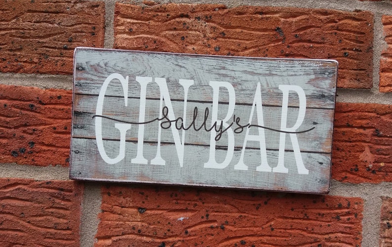 May include: A white wooden sign with the text "GIN BAR" in black lettering and "Sally's" written in cursive above the word "BAR". The sign has a distressed, weathered look.