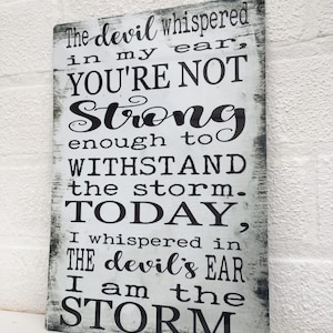May include: A white metal sign with black text that reads "The devil whispered in my ear, YOU'RE NOT Strong enough to WITHSTAND the storm. TODAY, I whispered in THE devil's EAR I am the STORM."