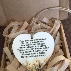 Op de afbeelding: Witte, hartvormige keramische hanger met een bloemenmotief en de tekst "You are my sunshine My only sunshine You make me happy When skies are grey You'll never know, dear How much I love you Please don't take My sunshine away"