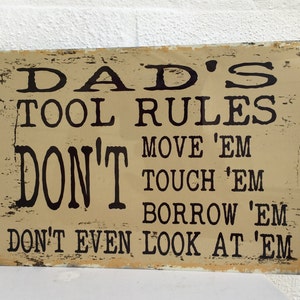 May include: A rustic-style sign with the text "DAD'S TOOL RULES DON'T MOVE 'EM TOUCH 'EM BORROW 'EM DON'T EVEN LOOK AT 'EM" in black lettering on a beige background.