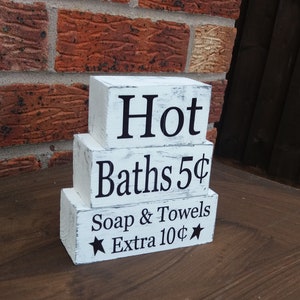 May include: White wooden blocks stacked in a pyramid shape. The top block reads "Hot", the middle block reads "Baths 5¢", and the bottom block reads "Soap & Towels Extra 10¢".