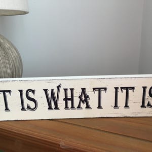 Può includere: Un cartello di legno bianco con lettere nere che recita "It is what it is".