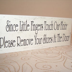 Op de afbeelding: Een wit houten bord met zwarte tekst die luidt: "Since little fingers touch our floor, please remove your shoes at the door."