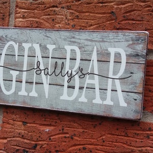 May include: A rustic wooden sign with the text "Gin Bar Sally's" in white lettering. The sign has a distressed, weathered look and is perfect for a bar or home decor.