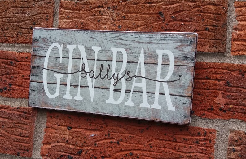 May include: A rustic wooden sign with the text "Gin Bar Sally's" in white lettering. The sign has a distressed, weathered look and is perfect for a bar or home decor.