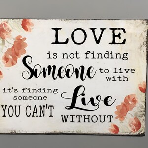 Puede incluir: Un letrero rectangular de madera con un fondo blanco desgastado. El letrero presenta las palabras "LOVE is not finding Someone to live with, it's finding someone YOU CAN'T Live WITHOUT" en escritura negra. Acentos florales de acuarela en tonos rojos y melocotón en las esquinas.