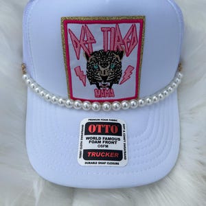 Może przedstawiać: Biała czapka trucker z naszywką w kolorze różowym i złotym przedstawiającą lamparta z napisem "Da Tired Mama". Czapka ma ozdobny pasek z pereł wokół ronda.