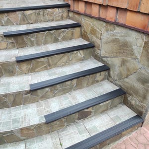 May include: A set of black non-slip treads for stairs. The treads are installed on a set of stone stairs with a brick wall in the background.