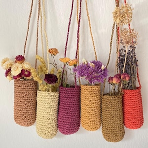May include: A collection of seven hanging crochet vases, each filled with dried flowers. The vases are in shades of brown, yellow, orange, and purple, and are suspended by cords. The flowers are in shades of red, yellow, purple, and white.