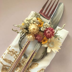 Peut inclure: Une présentation de table décorative avec une fourchette et un couteau, ornée d'une composition florale. L'argenterie repose sur une serviette blanche avec des broderies dorées. Les fleurs comprennent des fleurs jaunes, roses et crème, créant une esthétique élégante.