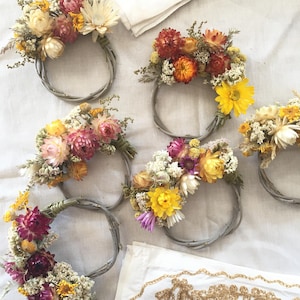 Pode incluir: Uma coleção de pequenas coroas de flores decorativas. Cada coroa é feita de uma base de galhos cinzentos e adornada com flores secas em tons de amarelo, rosa, laranja e branco. As coroas são dispostas sobre uma superfície branca.