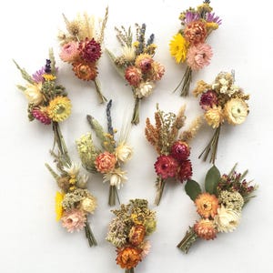 Può includere: Una collezione di dieci piccoli bouquet di fiori secchi. I bouquet presentano una varietà di colori, tra cui rosa, giallo, arancione e crema. Ogni bouquet è legato con spago, creando un'estetica rustica e naturale.