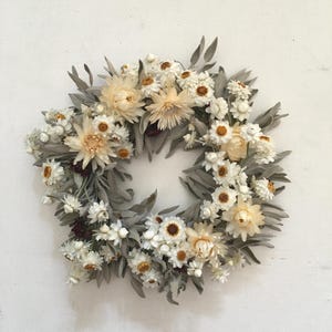 Op de afbeelding: Een ronde krans samengesteld uit gedroogde bloemen en bladeren. Het arrangement bevat crèmekleurige en witte strobloemen met gele harten, afgewisseld met lange, smalle, saliegroene bladeren en kleine donkerbruine accenten door het geheel.
