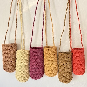 May include: Seven handmade crochet bottle holders in shades of brown, beige, purple, yellow, and red-orange. Each holder has a long strap for carrying and is designed to hold a bottle. The holders are suspended against a white wall.