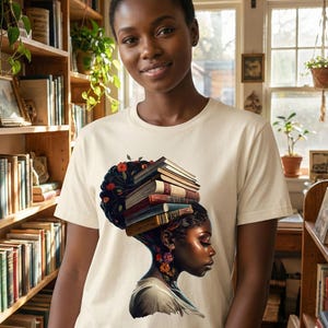 May include: A cream-colored t-shirt featuring a graphic of a woman with a stack of books on her head, adorned with flowers. The woman's profile is detailed with vibrant colors. The background includes bookshelves filled with books.