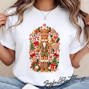 May include: A white t-shirt featuring a central graphic of a gingerbread nutcracker surrounded by holiday elements including candy canes, green Christmas trees, red bows, snowflakes, gingerbread cookies, and wrapped gift boxes at the base.