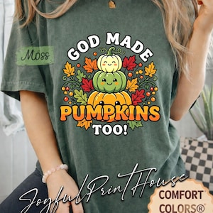 Op de afbeelding: Een mosgroen T-shirt met korte mouwen en een afbeelding van drie gestapelde pompoenen met lachende gezichten, omringd door herfstbladeren en bessen. De tekst "GOD MADE PUMPKINS TOO!" staat in dikke, witte en oranje letters boven en onder de pompoenen.