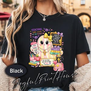 May include: A black t-shirt featuring a graphic of a pastel-striped ghost wearing a yellow bow and pearl necklace. The design includes books, a pencil cup, an apple, a backpack, and text reading "TEACHING little minds HIS GLORY" and Bible verses.