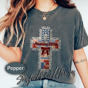 以下が含まれることがあります： 赤、白、青のパッチワークでデザインされた十字架が特徴のグレーのTシャツ。「America 250th」の文字と赤いリボンが付いています。カジュアルなスタイルです。