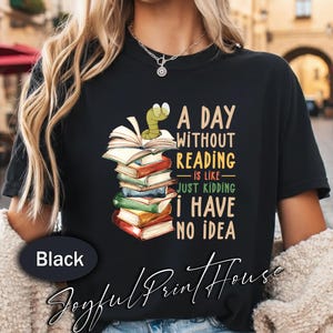 Pode incluir: Camiseta preta com o texto "A DAY WITHOUT READING is like - JUST KIDDING I HAVE NO IDEA." O design apresenta uma pilha de livros com um verme verde no topo. A marca "Joyful Print House" está na parte inferior.