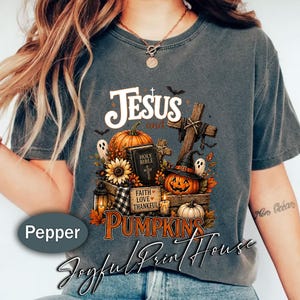 May include: A charcoal gray t-shirt featuring a graphic print with the text Jesus and Pumpkins, a Holy Bible, a wooden cross, pumpkins, sunflowers, ghosts, and a lantern. The design includes a small sign that reads Faith Love Thankful.