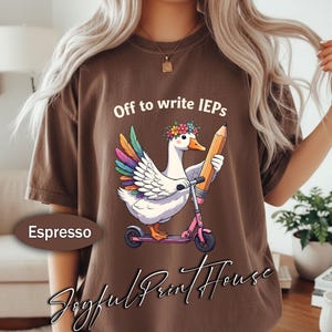Könnte beinhalten: Ein braunes T-Shirt mit einer Grafik einer weißen Gans mit regenbogenfarbenen Flügeln, die einen Blumenkranz trägt, auf einem rosa Roller fährt und einen großen gelben Stift hält. Der Text über der Gans lautet, Off to write IEPs.