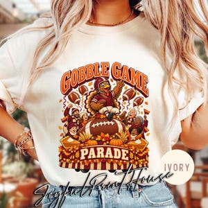 Könnte beinhalten: Ein elfenbeinfarbenes Sweatshirt mit Rundhalsausschnitt und einem Grafikdruck mit dem Text GOBBLE GAME PARADE. Das Design zeigt einen Cartoon-Truthahn in Football-Uniform, einen großen Football, Herbstblätter, Kürbisse, Torten und eine jubelnde Menge.