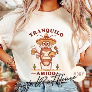 May include: Ivory-colored sweatshirt featuring a cartoon cockroach wearing a sombrero, holding a beer, a shot, a taco, and a maraca. The text "TRANQUILO" is above the cockroach, and "AMIGO" is below. The sweatshirt has the text "Joyful Print House" at the bottom.