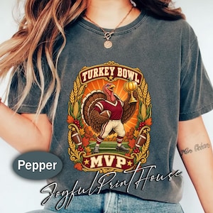 May include: A charcoal grey t-shirt featuring a graphic of a turkey dressed in a football uniform holding a football and a trophy. The text reads Turkey Bowl and MVP. The design is framed by wheat stalks, corn, and autumn leaves on a field.