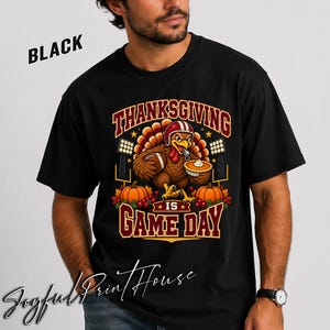 Könnte beinhalten: Ein schwarzes T-Shirt mit einer Grafik eines Truthahns, der einen Footballhelm trägt und einen Football sowie einen Kürbiskuchen hält. Der Text "THANKSGIVING IS GAME DAY" ist in fetten roten und gelben Buchstaben abgebildet.