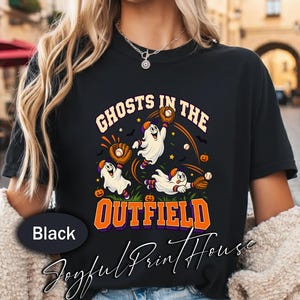 May include: A black t-shirt featuring a graphic of three white ghosts playing baseball. The ghosts wear baseball caps and gloves, catching baseballs. The text reads GHOSTS IN THE OUTFIELD in orange and purple letters, with small pumpkins and bats.