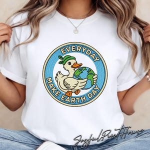 Puede incluir: Camiseta blanca con un gráfico circular de un pato con un sombrero verde y sosteniendo un globo terráqueo. El gráfico dice "EVERYDAY MAKE EARTH DAY" en azul y amarillo. La camiseta es de estilo casual.