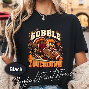 Könnte beinhalten: Ein schwarzes T-Shirt mit einer Grafik eines Truthahns, der einen Footballhelm und ein Trikot trägt und einen Football hält. Der Text lautet GOBBLE TOUCHDOWN in fetten orangefarbenen Buchstaben. Das Design enthält Herbstblätter und Beeren in Rot und Gold.
