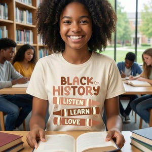 Könnte beinhalten: Ein beiges T-Shirt mit den Worten "BLACK HISTORY" in braunen Buchstaben und den Worten "LISTEN, LEARN, LOVE" in Buntstift-Grafiken. Eine junge Person lächelt, während sie ein offenes Buch hält. Andere Personen sind im Hintergrund.