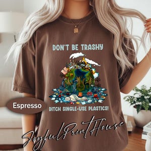 Puede incluir: Camiseta color espresso con el texto "DON'T BE TRASHY" sobre una ilustración de la Tierra contaminada. El gráfico incluye fábricas, basura y las palabras "DITCH SINGLE-USE PLASTICS!"
