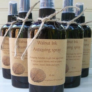 May include: Several dark brown glass spray bottles of "Walnut Ink Antiquing Spray" are arranged in a row. Each bottle has a tan label with text and a walnut image, tied with twine. The bottles are 4 oz.